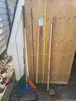 Full view of 6 gardening tools standing upright against a wooden background: 2 leaf rakes (blue and orange), a metal garden rake, hand cultivator, hoe, and weeding tool.