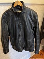 Front view of the black leather jacket hanging on a wooden hanger, showing full length with zip closure and pockets.