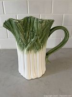 Front angled view of green onion shaped pitcher, showing detailed texture and green to white glazing, on countertop with tile backsplash