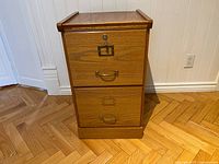 Front view of the small wooden filing cabinet with two drawers, brass handles, and a label holder on top drawer which features a lock.