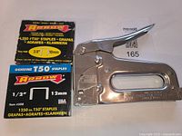 Photo showing the front view of the Arrow T-50m staple tool positioned beside two unopened boxes of Arrow genuine T-50 staples - one 3/8 inch size and one 1/2 inch size.
