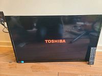 Toshiba 32-inch flat-screen television powered on showing Toshiba logo, with remote control resting on right side of TV on floor