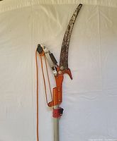 Full view of the tree pruner showing the curved saw blade, orange pruner body with rope mechanism and part of telescoping pole