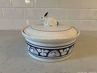 Side view of the covered casserole dish showing white crackle glaze, blue decorative band, and white bunny knob handle on lid.