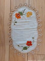 Top view of oval vintage doily showing floral embroidery with orange, red, and yellow flowers and green leaves on white fabric and beige lace trim around edges.