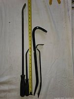 Six black metal wrecking and pry bars of various sizes and shapes laid out on a white cloth background with a tape measure showing lengths. Two pry bars have black handles, others are flat metal bars with bent or forked ends.
