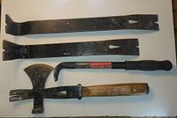 Four wrecking tools laid out on white surface: two flat pry bars, one wooden handle crate hammer with axe head, and one 11-inch nail puller with rubber handle.