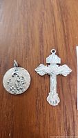 Top view of two sterling silver pendants on wood surface: a round religious medallion and an ornate crucifix pendant.