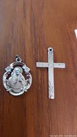 Two sterling silver pendants on a wooden surface: one oval religious Jesus pendant with floral designs, one flat cross pendant with engraved patterns.
