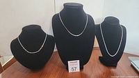 Three sterling silver chains displayed on black necklace stands on wooden surface with an auction lot card numbered 57