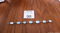 Full length view of the sterling silver bracelet with seven tear-drop shaped light blue stones laid flat on a wood surface with a lot number card nearby.