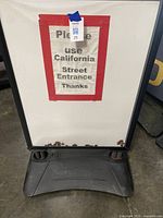 Close up of a black framed upright sign stand with a white sign taped inside bordered red stating 'Please use California Street Entrance Thanks' with a plastic weighted base that has a curved bottom and dual springs holding the frame.