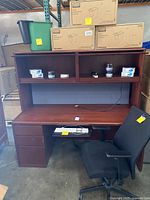 View of the wood office desk with top hutch filled with small items, keyboard on pull-out, and black mesh office chair.