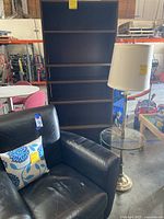 Black leather chair, tall dark brown bookshelf and glass floor lamp beside them in warehouse.