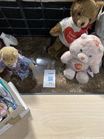 Photo showing brown bear with 'Bear Hugs' t-shirt, light pink poodle, and other stuffed animals on table.