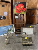 Photo of wood ladder shelving unit with decorative floral and landscape paintings leaning against it, glass top coffee table with brass curved legs in front, and metal book rack on floor.