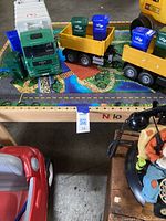 Photo showing toy trucks with attached trailers holding small blue recycling bins on a play rug with road print; part of ride-on toy car visible, along with a vintage Goofy rotary dial phone on a wooden box.