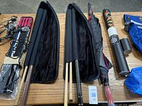 Photo showing four cue sticks on a wooden table, two in black cases, next to a black and red umbrella and blue bag.