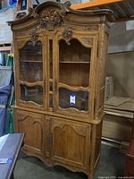 Full view of the ornate wooden china cabinet showing detailed hand-carved floral and scrollwork designs on upper and lower doors. Clear glass panels on upper doors, lower doors solid wood with carvings. Some wear visible.