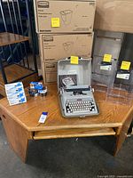 Corner oak desk with Royal Futura 600 gray manual typewriter on top, several Rubbermaid side bins stacked in boxes behind and small office supplies on the desk