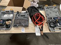 All items displayed on table including drill in open grey case, bit socket set, jumper cables coiled, and sander in case.