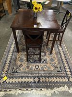 Square dark brown wood dining table with three matching chairs and artificial flower bouquet on top, placed over a beige and navy patterned rug.