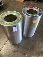 Two cylindrical metal trash bins labeled 'Compost' (green rim) and 'Recycle' (blue rim) standing on concrete floor.