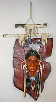 Front view of Indigenous wall hanging showing carved face with feathers and ribbons suspended by wooden rods.