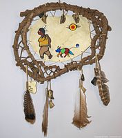 Full view of Indigenous wall hanging with painted rawhide, wooden frame, hanging deer hooves and feathers.