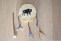 Front view of the small native drum showing black painted buffalo on the deer hide drum head, leather cords with beads and feathers hanging on the side, and wooden drum beater beside the drum.