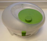 Top angled view of salad spinner with clear plastic lid, green rotary handle, and green clip.
