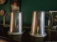 Two Sheffield pewter beer steins on a wood surface. Both have solid handles and cylindrical bodies with polished silver-tone finish. Left stein engraved with Stewarton Bonnet Guild.