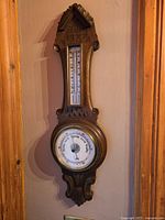 Full view of carved wooden wall-mounted barometer and thermometer with brass bezel and visible scales and dial.