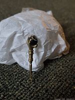 Close-up photo showing a vintage brass button hook with claw holding a dark glass marble, resting on a carpeted surface with a white crumpled paper in the background.
