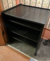 Front and side view of a black computer table with two shelves and castors, against floral wallpaper and wooden floor.