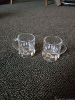 Two small clear glass beer mugs photographed on carpeted floor to show condition and size.
