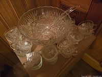 Glass punch bowl set with 14 matching cups, one plastic ladle, and several metal hangers displayed on wooden surface.