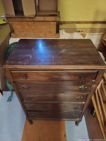 Tall hardwood chest with five drawers, top surface with light marks, standing on turned legs in a room with other furniture.