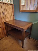 Wooden square end table placed against wall with wood paneling and mirror above. Shows side and top surfaces, tapered legs, and lower shelf in medium brown finish.