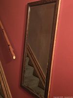 Photo showing a vertically mounted rectangular mirror with an oak wood frame and bevelled edges on the glass, against a dark red wall and near wooden banister.