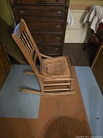 Side view of small wooden rocking chair showing curved rockers, turned spindle backrest, and damaged cane seat needing recaning.