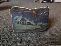 View of small rectangular dark fabric pillow with detailed embroidery of Portland Head Light lighthouse and keeper's house, with yellow text and gold braided edges.