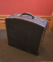 Photo of a vintage suitcase placed upright on carpet against a reddish wall, showing the front and top surfaces including handle and clasps
