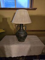 Full view of the Spanish pottery table lamp with pleated shade on a covered surface, showing its shape, handles, decorative patterns, and lampshade.