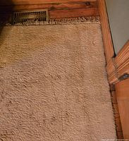 Close-up photo of the corner of a beige wool carpet with medium pile, showing some damage to the fringe and wooden floor vents nearby.