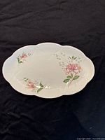 Top-down view of a scalloped oval white bone china dish decorated with pink honeysuckle flowers and embossed edge details on a black cloth.