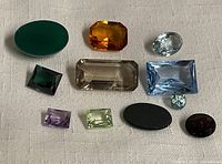 All 11 unmounted faceted and smooth natural gemstones in varied colors and cuts are displayed on white fabric background