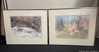 Two framed watercolour paintings side by side on black surface; left depicts snowy winter scene, right depicts autumn forest