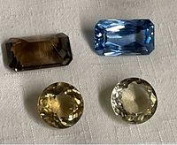 Four natural faceted loose gemstones including one dark brown rectangular Smokey Quartz, one blue rectangular Aquamarine, and two round light yellow-green stones photographed on a white cloth background.