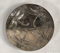 Top-down image showing round silver plated dish with four bas-relief dragonfly figures and tarnished surface.